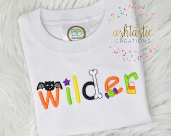May include: White t-shirt with a Halloween-themed embroidered name "Wilder". The letters are decorated with a bat, a star, a bone, candy, and a candy corn.