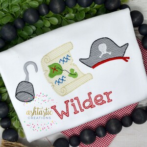 May include: White embroidered baby onesie with a red pirate hat, a treasure map, and a hook. The name "Wilder" is embroidered in red below the design.