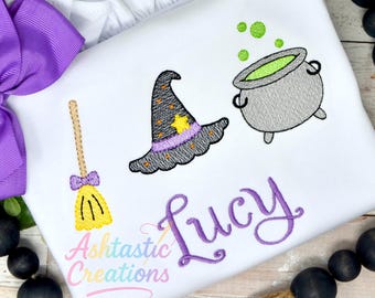 May include: White shirt with embroidered Halloween-themed designs. Features a witch's hat, cauldron with green bubbles, and a broom. The name "Lucy" is embroidered in purple script. The shirt also has the text "Ashtastic Creations".