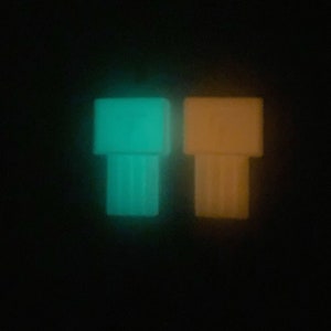 Puede incluir: Dos pequeñas figuritas de plástico que brillan en la oscuridad. Una es verde turquesa y la otra es naranja.