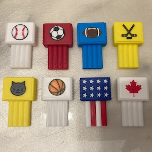 Puede incluir: Ocho bloques de plástico coloridos con diferentes imágenes en cada lado. Las imágenes incluyen una pelota de béisbol, una pelota de fútbol, una pelota de fútbol americano, palos de hockey, un gato, una pelota de baloncesto, la bandera estadounidense y la bandera canadiense.