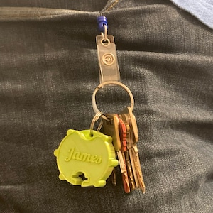 Puede incluir: Un llavero verde con el nombre "James" en él, unido a un juego de llaves con un llavero de plástico transparente y un clip azul.