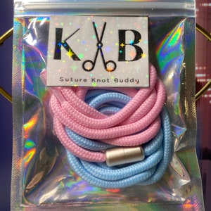 Puede incluir: Un nudo de sutura rosa y azul con una cuenta de metal plateada. El paquete tiene una etiqueta que dice "K&B Suture Knot Buddy".