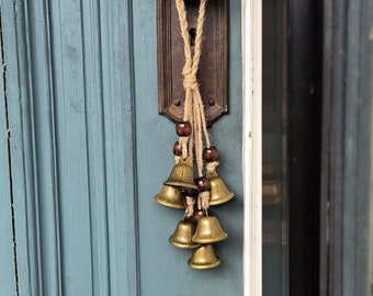 May include: A set of five small brass bells hanging from a door knob with brown wooden beads and twine. The bells are attached to a door with a blue painted surface.