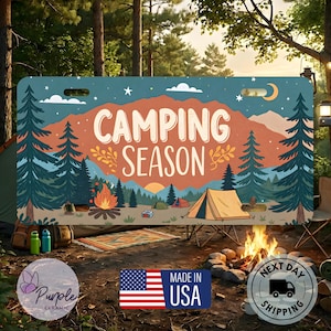 May include: A rectangular metal license plate featuring a graphic illustration of a forest landscape with mountains, pine trees, a campfire, and a tent. The text CAMPING SEASON is printed in white in the center. Two mounting slots are at the top.