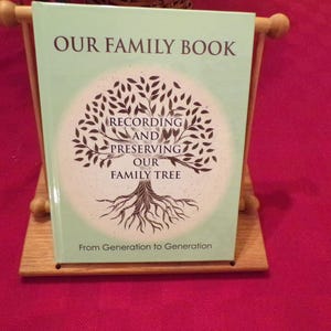 K&ouml;nnte beinhalten: Ein gr&uuml;nes Buch mit dem Titel "Our Family Book" und einer Baumillustration. Der Text unter dem Baum lautet "Recording and Preserving Our Family Tree" und "From Generation to Generation".
