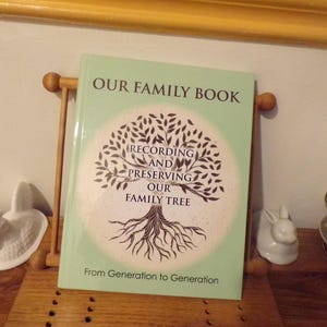 K&ouml;nnte beinhalten: Ein gr&uuml;nes Buch mit dem Titel "Our Family Book: Recording and Preserving Our Family Tree". Der Umschlag zeigt einen stilisierten Baum mit Wurzeln und &Auml;sten. Der Text "From Generation to Generation" ist unter dem Baum gedruckt.