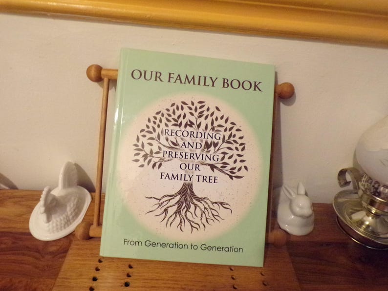 K&ouml;nnte beinhalten: Ein gr&uuml;nes Buch mit dem Titel "Our Family Book: Recording and Preserving Our Family Tree". Der Umschlag zeigt einen stilisierten Baum mit Wurzeln und &Auml;sten. Der Text "From Generation to Generation" ist unter dem Baum gedruckt.