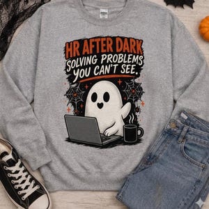 May include: A heather gray crewneck sweatshirt featuring a graphic of a white ghost sitting at a laptop with a black coffee mug. The text reads HR AFTER DARK SOLVING PROBLEMS YOU CAN'T SEE, surrounded by black spiderwebs and small orange stars.