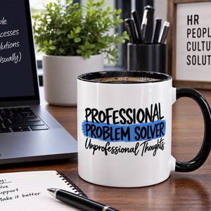 May include: A white ceramic mug with a black handle and interior rim. The front features the black text "PROFESSIONAL PROBLEM SOLVER" over a blue brushstroke graphic, with the black script text "Unprofessional Thoughts" printed underneath.