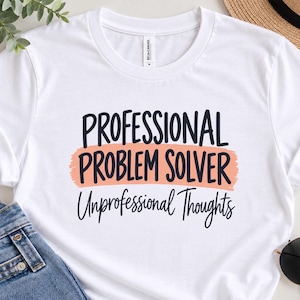 May include: A white short-sleeved t-shirt featuring the black text "PROFESSIONAL PROBLEM SOLVER" over a peach-coloured brushstroke graphic, with the black text "Unprofessional Thoughts" printed in a cursive script font underneath the graphic.