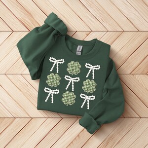 May include: A dark green sweatshirt featuring a pattern of white bows and green gingham shamrocks. The long sleeves are cuffed. The design is centered on the front of the sweatshirt, creating a festive St. Patrick's Day theme.