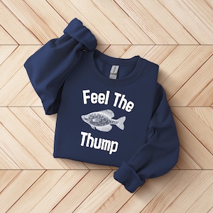 May include: Navy blue sweatshirt with the text "Feel The Thump" and a white fish graphic. The sweatshirt is folded on a wooden surface.