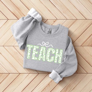 Pode incluir: Um moletom cinza com a palavra "TEACH" em verde, com um padrão de trevos. As mangas estão dobradas, revelando o interior branco. Um laço decorativo está acima da palavra "TEACH". O moletom está sobre uma superfície de madeira.