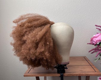 Op de afbeelding: Een lichtbruine, krullende en getextureerde pruik wordt getoond op een beige canvas etalagepophoofd. De pruik heeft volumineuze, strakke krullen die naar buiten uitsteken. Het hoofd is gemonteerd op een zwarte verstelbare metalen standaard.