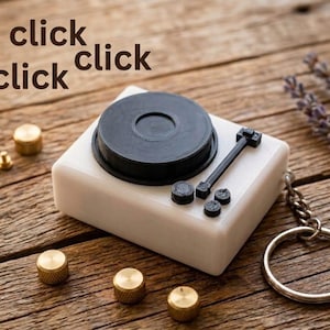Può includere: Un portachiavi con giradischi in miniatura bianco con un disco nero e un braccio. Il portachiavi ha un anello e una catena argentati. L'immagine mostra anche tre manopole color oro e il testo "click click click".