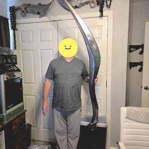May include: A large, curved, metallic blade with a pointed tip and a handle at the base. The blade features a polished, reflective silver surface that tapers along its length. It is held vertically, showcasing its significant height and slender, sweeping design.