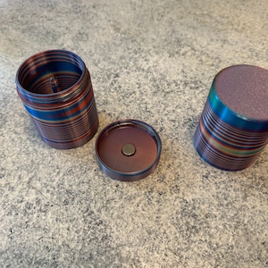 May include: A three-piece, cylindrical grinder with a vibrant, multi-colored finish. The grinder is disassembled, showing the base, lid, and the main body. The colors include blue, red, and gold, creating a striped pattern.