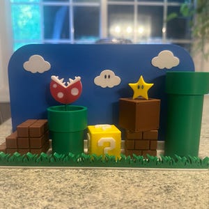 May include: A colorful 3D-printed diorama featuring elements from the Super Mario video game. It includes a blue backdrop with white clouds, a red and white Piranha Plant, green pipes, a yellow question mark block, and a yellow star.