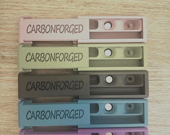 Peut inclure: Cinq étuis en plastique de couleurs différentes avec le texte "CARBONFORGED" imprimé sur le devant. Les étuis sont disposés verticalement, l'étui rose en haut et l'étui violet en bas.