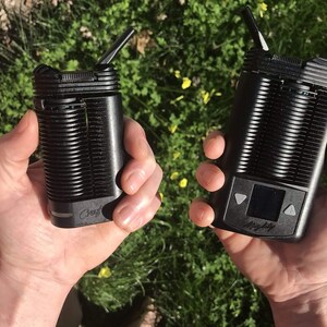 May include: Two black vaporizers are held in hands against a background of green grass and yellow flowers. One device has a mouthpiece extended, while the other features a small screen and control buttons. The word "Mighty" is visible on one device.