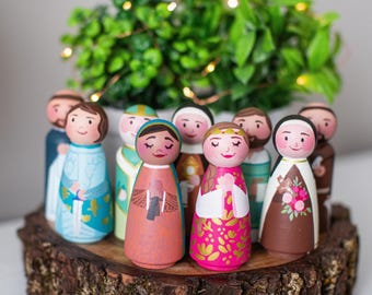 May include: A collection of hand-painted wooden peg dolls, each depicting a different figure, arranged on a rustic wooden base. The dolls feature detailed clothing and facial expressions, with a variety of colors and designs. A green plant and fairy lights are in the background.