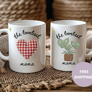 May include: Two white ceramic mugs with the text "the loveliest mama". One mug features a red gingham heart, the other a green gingham plant in a brown pot. Both mugs have a white handle and yellow star accents.