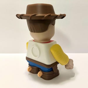 Pu&ograve; includere: Figura giocattolo stampata in 3D di un personaggio cowboy, vista da dietro. La figura indossa un cappello da cowboy marrone, una camicia bianca con colletto rosso, pantaloni blu e stivali marroni con speroni gialli. Le braccia sono gialle.