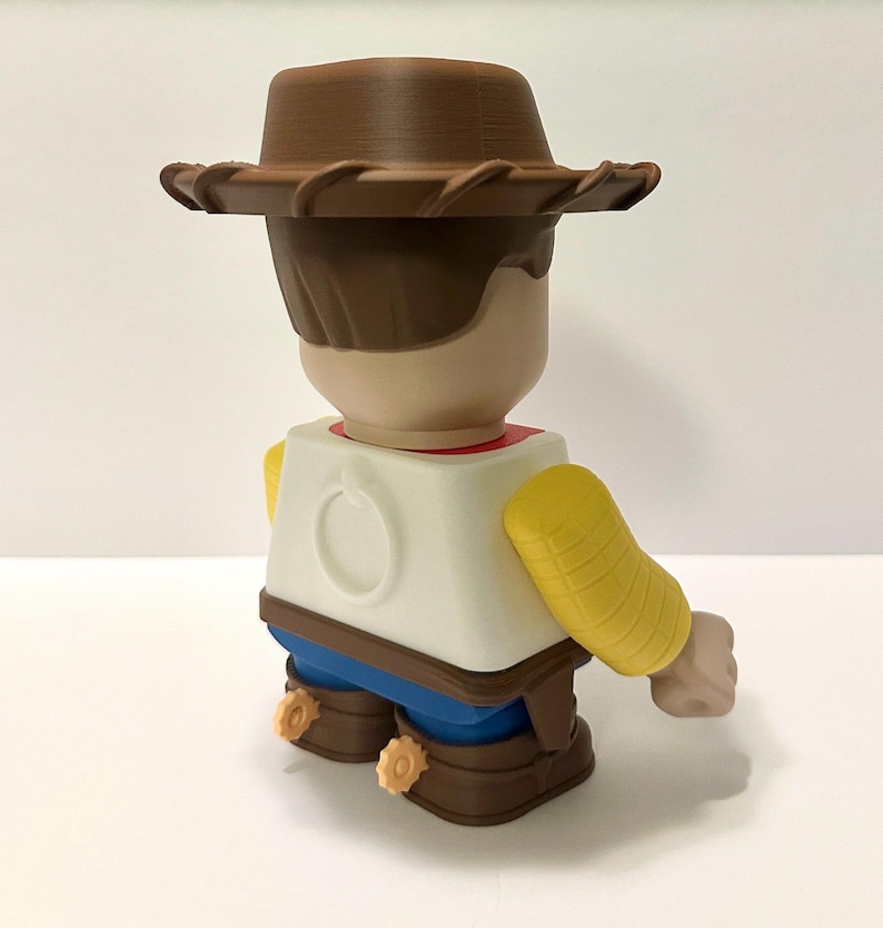 Pu&ograve; includere: Figura giocattolo stampata in 3D di un personaggio cowboy, vista da dietro. La figura indossa un cappello da cowboy marrone, una camicia bianca con colletto rosso, pantaloni blu e stivali marroni con speroni gialli. Le braccia sono gialle.