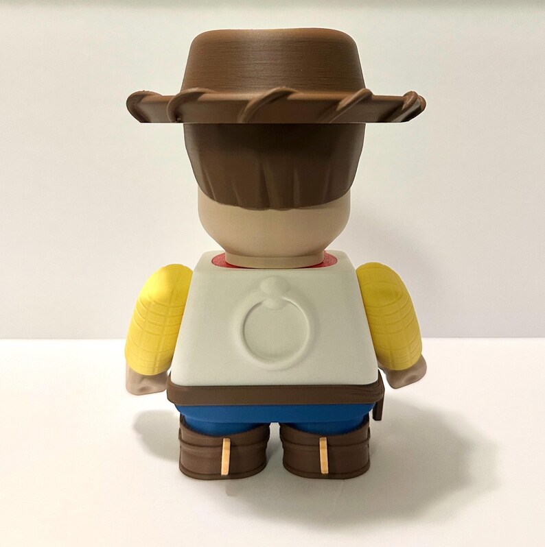 Pu&ograve; includere: Una statuetta da collezione di un personaggio dei cartoni animati di un cowboy, vista da dietro. La statuetta indossa un cappello da cowboy marrone, una camicia bianca con un disegno circolare, pantaloni blu e stivali marroni. Accenti gialli sono sulle spalle.