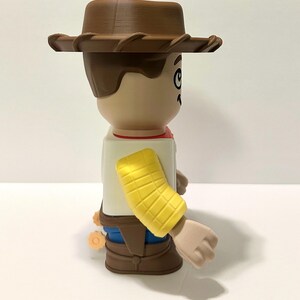 Pu&ograve; includere: Statuetta di un personaggio dei cartoni animati di un cowboy, con cappello marrone, capelli castani e viso abbronzato. La figura indossa una camicia bianca, un fazzoletto rosso, pantaloni blu e stivali marroni. &Egrave; visibile un bracciale giallo.