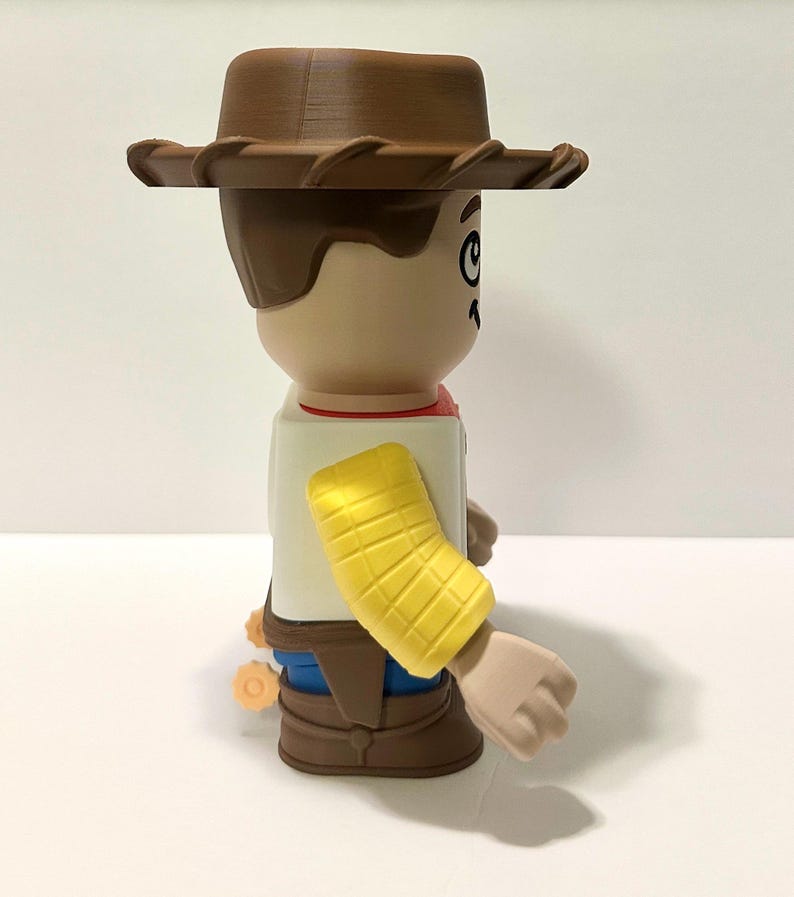 Pu&ograve; includere: Statuetta di un personaggio dei cartoni animati di un cowboy, con cappello marrone, capelli castani e viso abbronzato. La figura indossa una camicia bianca, un fazzoletto rosso, pantaloni blu e stivali marroni. &Egrave; visibile un bracciale giallo.