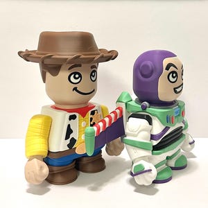 May include: Two stylized toy figures: Woody, in a brown hat, yellow shirt, and blue jeans, and Buzz Lightyear, in a white and green spacesuit with a purple helmet. Both figures have large, smiling faces.