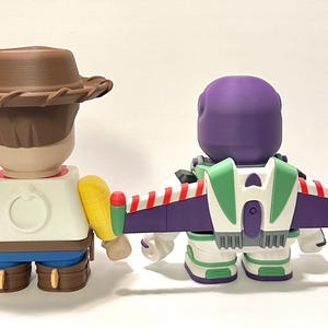 May include: Two collectible figurines, one resembling a cowboy with a brown hat and the other a space ranger with a purple helmet and green and white accents. The space ranger has a purple and white wing pack with red stripes.
