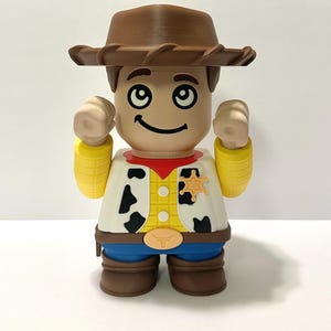 Pu&ograve; includere: Figurina giocattolo stampata in 3D di un personaggio cowboy, con cappello marrone, distintivo da sceriffo e gilet con macchie nere di mucca. La figura ha un volto sorridente e le braccia alzate.