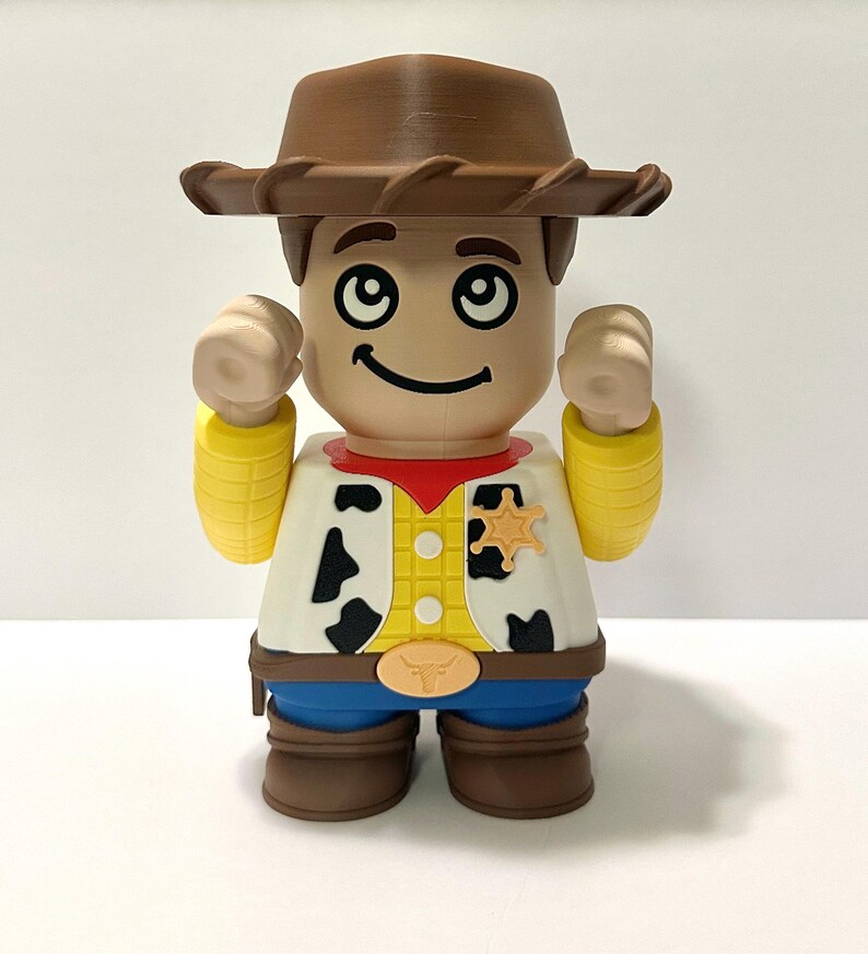 Pu&ograve; includere: Figurina giocattolo stampata in 3D di un personaggio cowboy, con cappello marrone, distintivo da sceriffo e gilet con macchie nere di mucca. La figura ha un volto sorridente e le braccia alzate.
