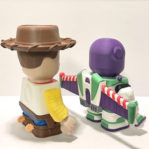 May include: Two 3D-printed toy figures: Woody, a cowboy with a brown hat, and Buzz Lightyear, a space ranger with a purple helmet and green and white wings. The toys are colorful and detailed, perfect for collectors or fans of the movie.