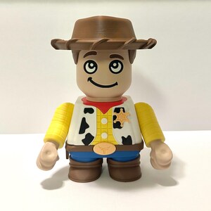 Pu&ograve; includere: Statuetta di un personaggio cowboy, con cappello marrone, gilet bianco con macchie nere, camicia gialla, pantaloni blu e stivali marroni. Il personaggio ha un distintivo da sceriffo e un volto sorridente.