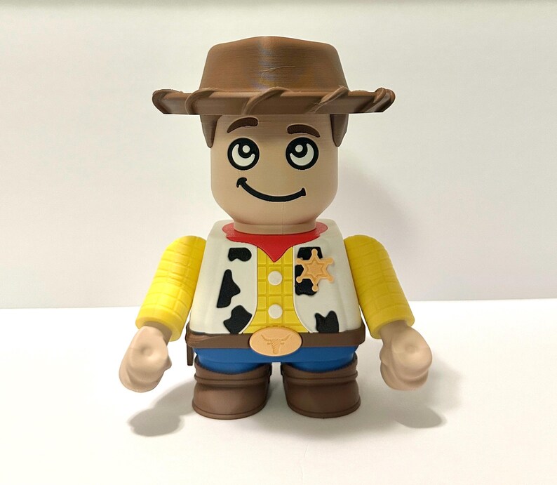 Pu&ograve; includere: Statuetta di un personaggio cowboy, con cappello marrone, gilet bianco con macchie nere, camicia gialla, pantaloni blu e stivali marroni. Il personaggio ha un distintivo da sceriffo e un volto sorridente.