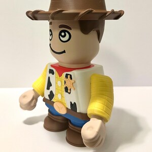 Pu&ograve; includere: Una statuetta giocattolo di un cowboy con cappello marrone, viso abbronzato e camicia gialla con macchie nere. La statuetta ha un fazzoletto rosso, pantaloni blu e stivali marroni. Un distintivo da sceriffo &egrave; visibile sul gilet.