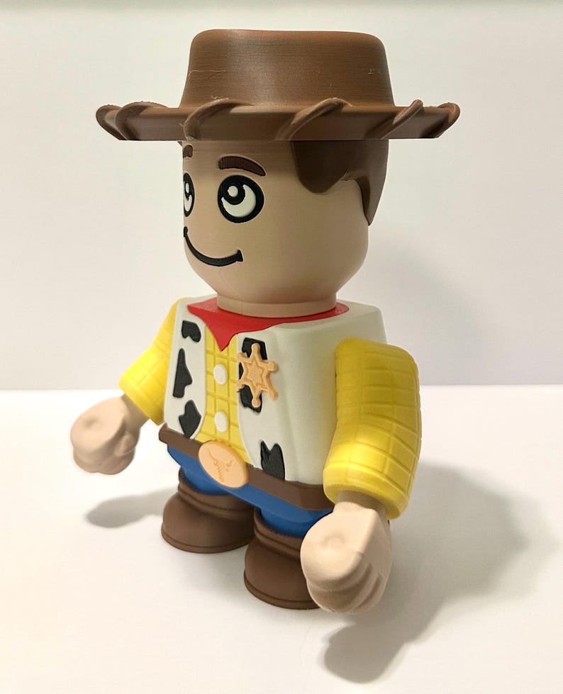 Pu&ograve; includere: Una statuetta giocattolo di un cowboy con cappello marrone, viso abbronzato e camicia gialla con macchie nere. La statuetta ha un fazzoletto rosso, pantaloni blu e stivali marroni. Un distintivo da sceriffo &egrave; visibile sul gilet.