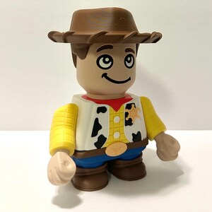 Pu&ograve; includere: Una statuetta di un cowboy con cappello, capelli e stivali marroni. La statuetta ha un viso abbronzato, un gilet bianco con macchie nere, una camicia gialla e pantaloni blu. Un distintivo da sceriffo &egrave; sul gilet.