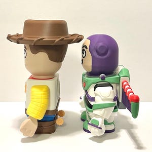 May include: Two stylized figurines of Woody and Buzz Lightyear from Toy Story. Woody wears a brown cowboy hat, a yellow sleeve, and a sheriff's badge. Buzz Lightyear has a purple helmet, green and white spacesuit, and red and white wings.