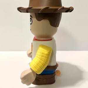 Pu&ograve; includere: Statuetta stilizzata di un personaggio cowboy, con cappello, capelli e stivali marroni. La figura ha un viso abbronzato, una manica gialla e un fazzoletto rosso. La figura indossa una camicia bianca e pantaloni blu.