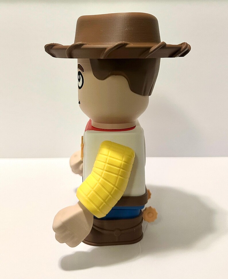 Pu&ograve; includere: Statuetta stilizzata di un personaggio cowboy, con cappello, capelli e stivali marroni. La figura ha un viso abbronzato, una manica gialla e un fazzoletto rosso. La figura indossa una camicia bianca e pantaloni blu.