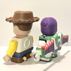 May include: Two collectible figurines: Woody in a brown cowboy hat, yellow shirt, and blue pants, and Buzz Lightyear in a green, white, and purple spacesuit. The toys are from the Toy Story franchise.