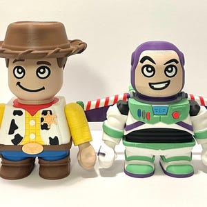 May include: Two collectible figures: Woody, a cowboy with a brown hat, yellow shirt, and blue jeans, and Buzz Lightyear, a space ranger in a green and white suit with purple accents. Both figures have large black eyes and smiling faces.
