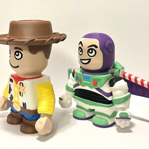 May include: Two collectible figures: Woody, a cowboy with a brown hat, yellow shirt, and blue pants, and Buzz Lightyear, an astronaut in a green and white spacesuit with a purple helmet and wings. Both figures have cartoonish features and are standing side-by-side.