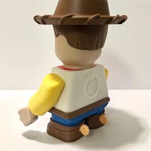 Pu&ograve; includere: Statuetta stilizzata di un personaggio cowboy, vista da dietro. La figura ha un cappello marrone, capelli castani e un viso abbronzato. Indossa una camicia bianca, pantaloni blu e stivali marroni. Le braccia sono gialle e la figura ha un disegno circolare sul retro.