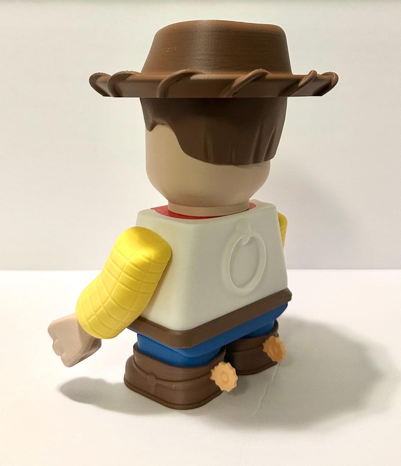 Pu&ograve; includere: Statuetta stilizzata di un personaggio cowboy, vista da dietro. La figura ha un cappello marrone, capelli castani e un viso abbronzato. Indossa una camicia bianca, pantaloni blu e stivali marroni. Le braccia sono gialle e la figura ha un disegno circolare sul retro.